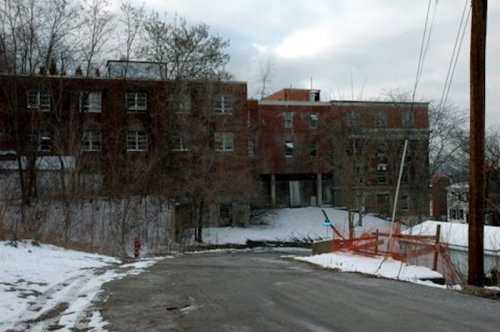 Abandoned building on a snowy road, surrounded by trees and construction barriers, under a cloudy sky.