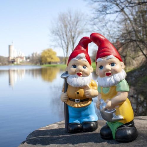 Two colorful garden gnomes stand by a serene lake, surrounded by trees and a distant building.