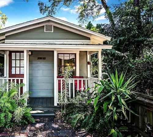 A charming green house with a porch, surrounded by lush greenery and plants.