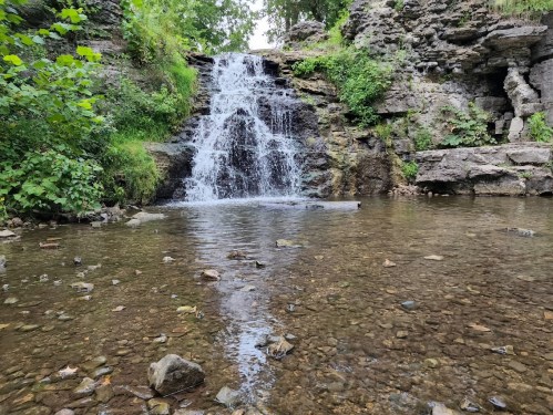 A serene waterfall cascades over rocks into a clear pool, surrounded by lush greenery and pebbles.