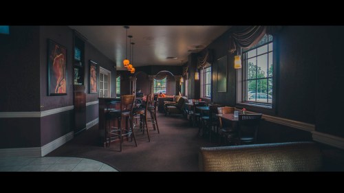 A cozy restaurant interior with dim lighting, tables, and bar seating, featuring large windows and decorative artwork.