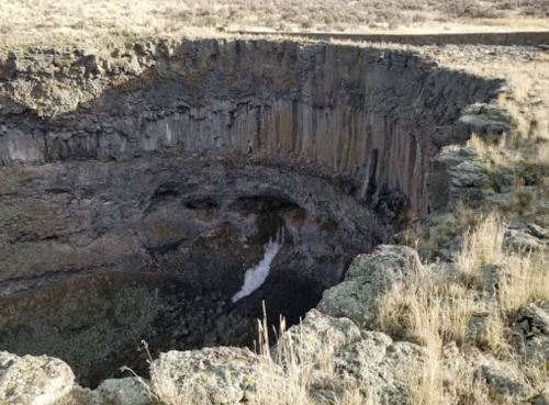 A deep, rocky crater with a small waterfall at the bottom, surrounded by dry grass and rugged terrain.