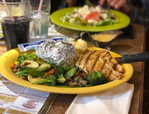 A plate with grilled fish, a baked potato, mixed vegetables, and a side of sauce, with drinks in the background.