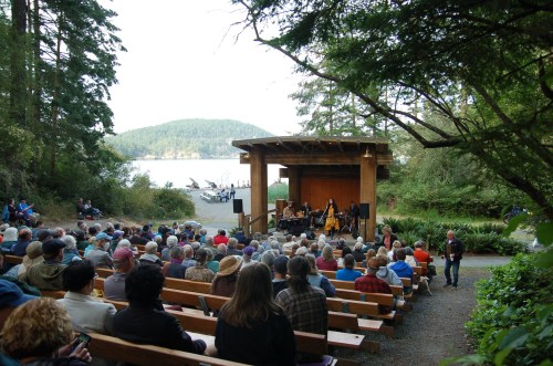 A crowd gathers for a performance at an outdoor stage by the water, surrounded by trees and hills.