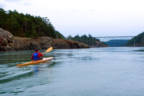 A person paddles a kayak through calm waters, with rocky shores and a bridge in the background.