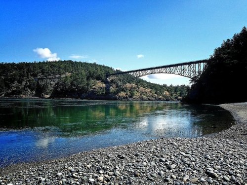 A scenic view of a bridge spanning over a calm river, surrounded by lush greenery and a clear blue sky.