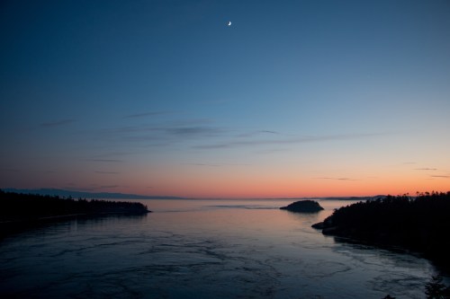 A serene twilight scene over a calm river, with a crescent moon and soft hues of orange and blue in the sky.