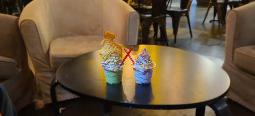 Two colorful ice cream desserts on a black table, one topped with a fish-shaped cone and sprinkles. Cozy café setting.