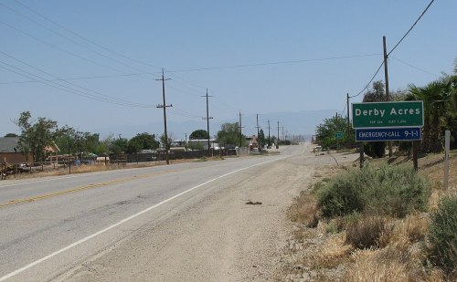 A rural road sign for Derby Acres, with power lines and sparse vegetation along the roadside on a sunny day.