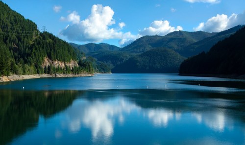 A serene lake surrounded by lush green mountains and blue skies, reflecting clouds on the water's surface.