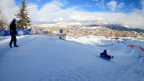 A child sleds down a snowy hill while an adult watches from above, with mountains and a town in the background.