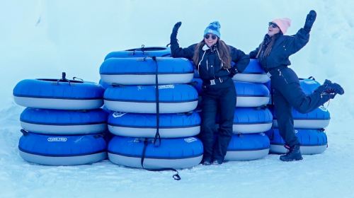 Two women in winter gear pose playfully beside a stack of blue snow tubes on a snowy landscape.