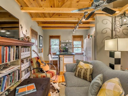 Cozy living space with wooden beams, a guitar, bookshelves, and a small kitchen area. Warm and inviting atmosphere.