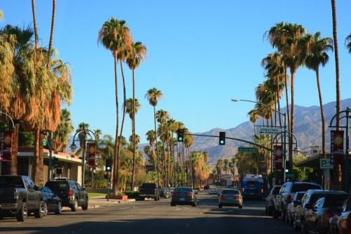 A sunny street lined with palm trees, cars parked along the road, and mountains in the background under a clear blue sky.