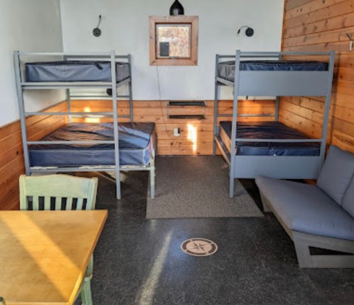 A simple room with two sets of bunk beds, a table, and a couch, featuring wooden walls and a small window.