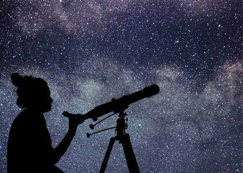 A silhouette of a person using a telescope against a starry night sky filled with stars and the Milky Way.