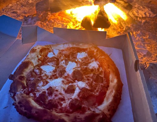 A pizza in a box sits in front of a glowing campfire, with warm colors reflecting off the crust.