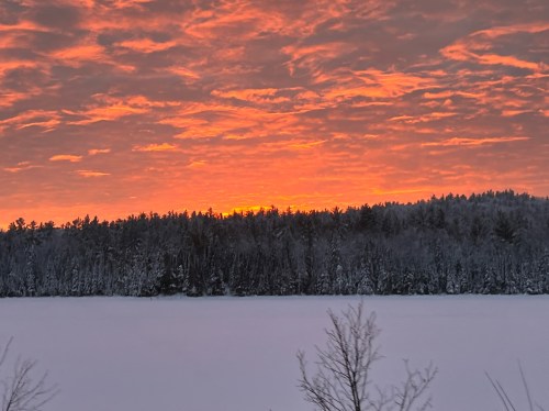 A vibrant orange sunset over a snowy landscape, with silhouetted trees against a colorful sky.