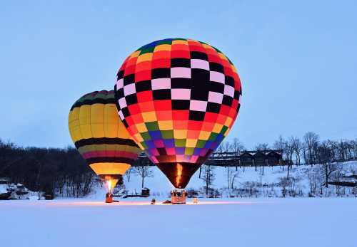 Two colorful hot air balloons glow against a snowy landscape at dusk, with trees and a building in the background.