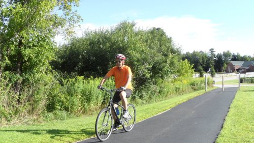 A person in an orange shirt rides a bicycle on a paved path surrounded by greenery and trees.