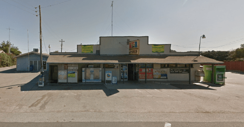 A small, weathered storefront with various signs and vending machines, set in a rural area under a clear sky.