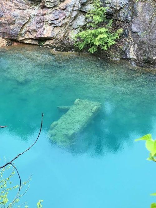 A submerged vehicle covered in moss, resting on the bottom of a clear blue-green body of water surrounded by rocky terrain.