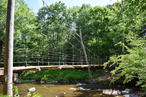 A wooden suspension bridge spans a calm stream, surrounded by lush green trees and a clear blue sky.