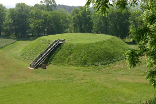 A grassy, circular earth mound with a wooden staircase leading up, surrounded by trees and open fields.