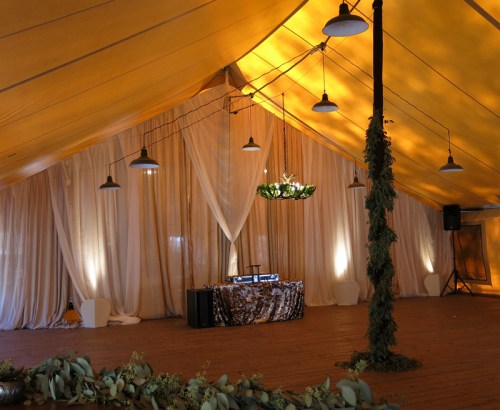 A spacious tent interior with warm lighting, draped fabric, and a DJ setup at the front, surrounded by greenery.