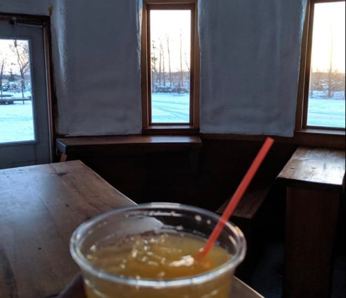 A hand holding a drink with a straw, with a view of snowy outdoors through large windows in a cozy setting.