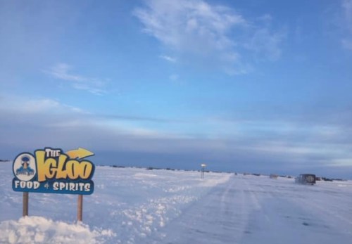 Sign for "The Igloo Food & Spirits" on a snowy road under a blue sky, with distant vehicles and snow-covered landscape.