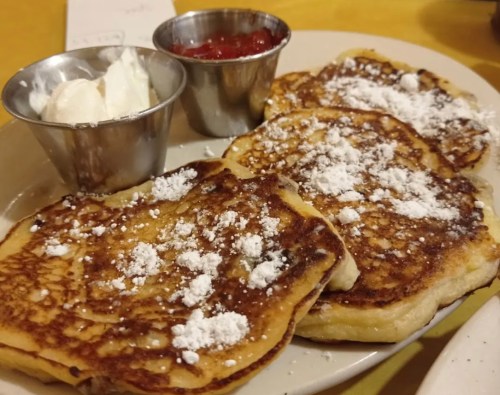 Three fluffy pancakes topped with powdered sugar, served with small cups of whipped cream and strawberry sauce.