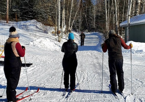 Three people in winter gear stand on a snowy path, preparing to ski, with trees and a cabin in the background.