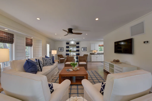 Bright living room with white sofas, a wooden coffee table, and a view of a kitchen, featuring modern decor and natural light.