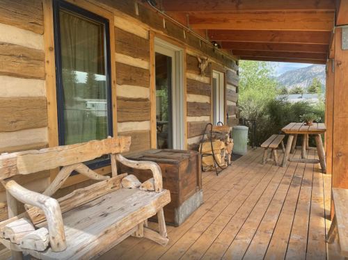 A rustic wooden porch with log walls, featuring a bench, a storage box, and a picnic table surrounded by greenery.