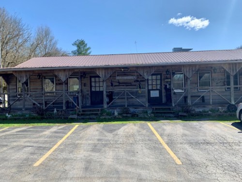 A rustic wooden building with a metal roof, surrounded by trees and a parking lot under a clear blue sky.