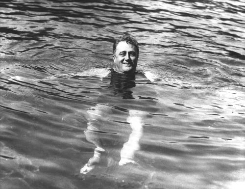 A smiling man swimming in a pool, partially submerged in water, with sunlight reflecting on the surface.