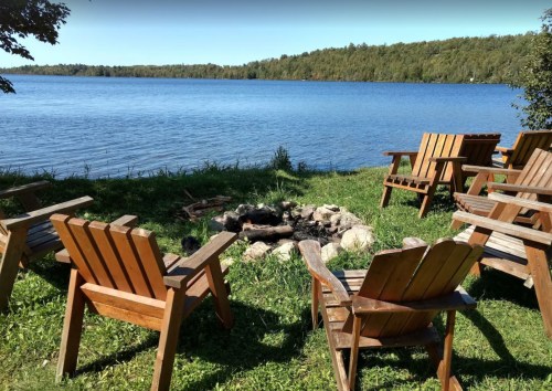 Wooden chairs arranged around a fire pit by a serene lake, surrounded by lush greenery and clear blue water.