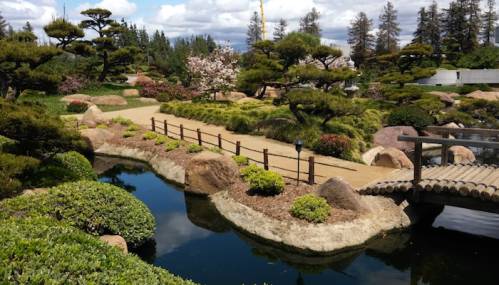 A serene Japanese garden with a winding path, pond, bridge, and lush greenery under a partly cloudy sky.