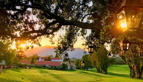 A scenic golf course at sunset, framed by trees and lanterns, with mountains in the background.