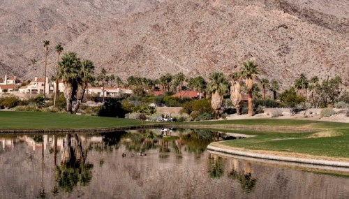 A serene golf course with palm trees and mountains reflected in a calm pond under clear skies.