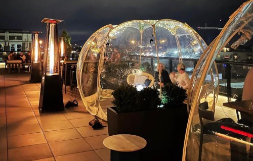 A rooftop dining area with transparent domes, heaters, and plants, illuminated at night with city lights in the background.