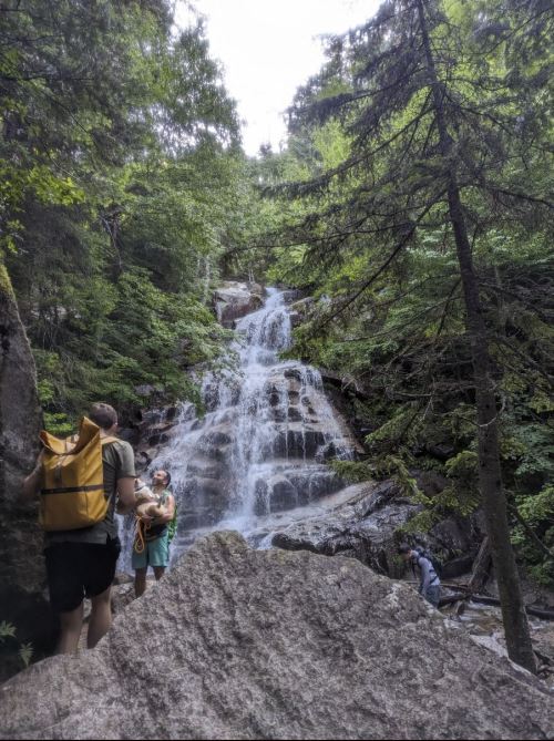 A waterfall cascades down rocky terrain, surrounded by lush greenery, with hikers exploring the area nearby.