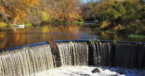 A serene waterfall cascades into a calm river, surrounded by vibrant autumn foliage.