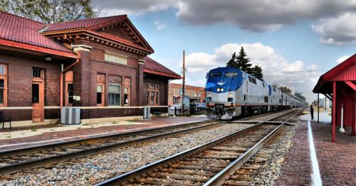 A train approaches a historic brick train station under a cloudy sky, with tracks and a red-roofed shelter nearby.