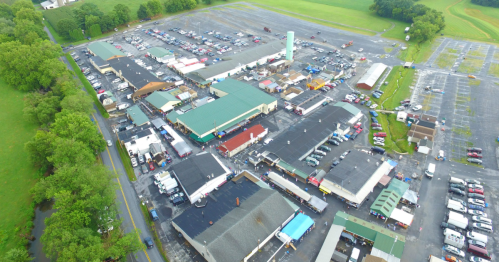 Aerial view of a large outdoor market with numerous stalls and parked cars, surrounded by green fields.