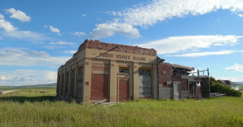 Abandoned building of First State Bank, partially collapsed, surrounded by tall grass under a blue sky with clouds.