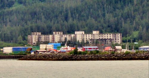 Abandoned buildings loom over a small harbor with colorful boats and containers, surrounded by lush green hills.