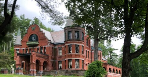 A large, red brick mansion with turrets, surrounded by trees and greenery under a cloudy sky.