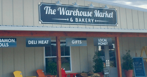 Sign for "The Warehouse Market & Bakery" with offerings like deli meat, gifts, and local honey, featuring colorful chairs outside.